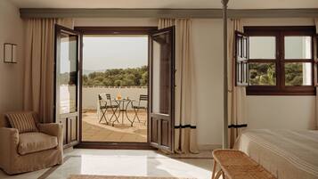 Un dormitorio con cama, un banco, una silla, una mesa con un vaso, y vistas a una terraza con mesa y sillas.