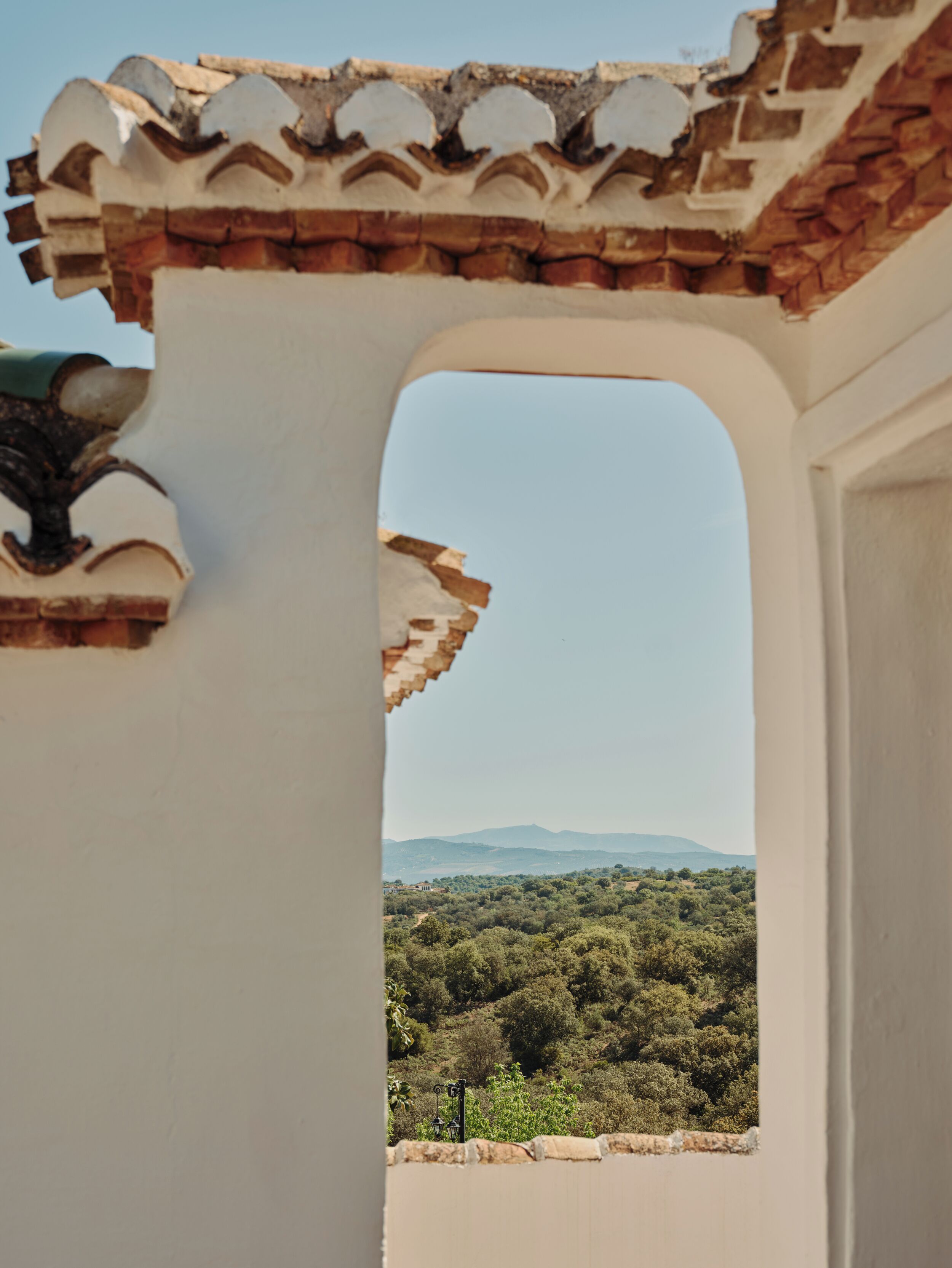 Vista a través de una ventana que da a un paisaje verde con árboles y montañas distantes, con una pared blanca y un tejado de tejas.