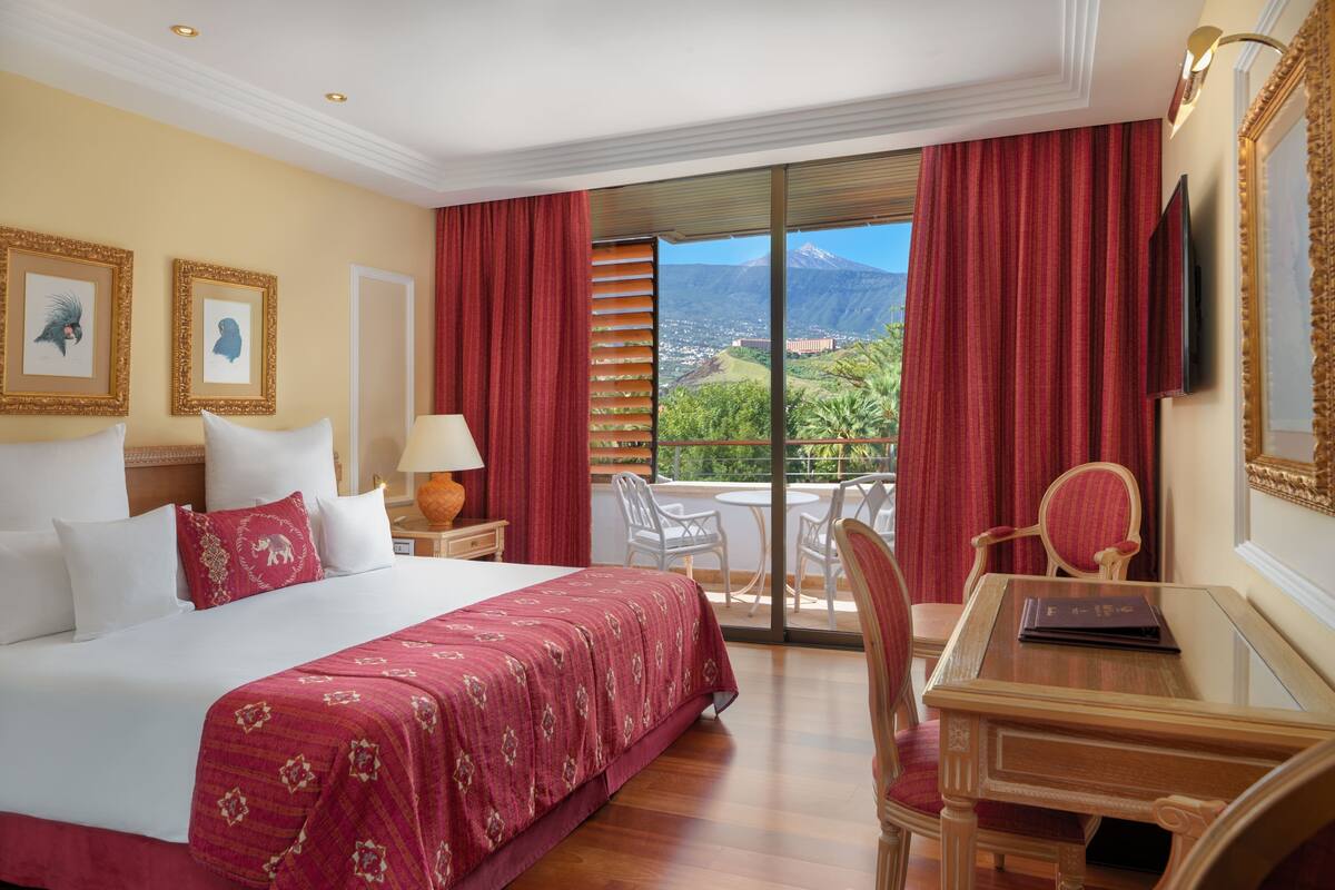 A hotel room with a large bed, a desk with a chair, a TV, and a balcony with a view of a mountain.