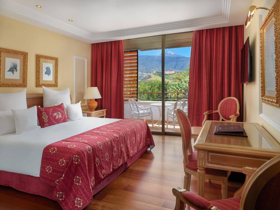 A hotel room with a large bed, a desk with a chair, a TV, and a balcony with a view of a mountain.
