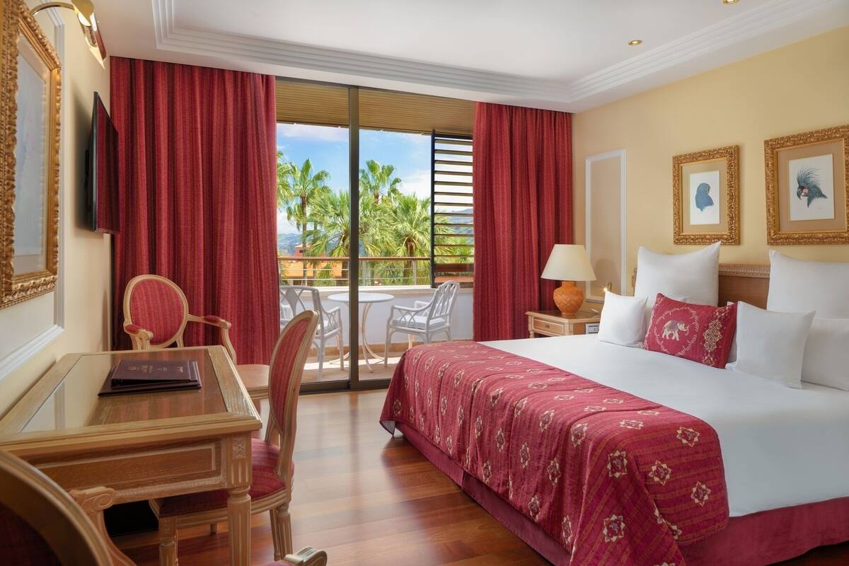 A hotel room with a large bed, a desk with a chair, a TV, and a balcony with red curtains.