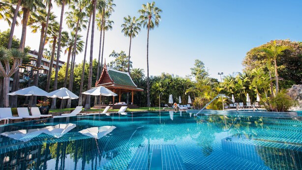 A swimming pool with tiled flooring, surrounded by lounge chairs and umbrellas, and palm trees in the background.