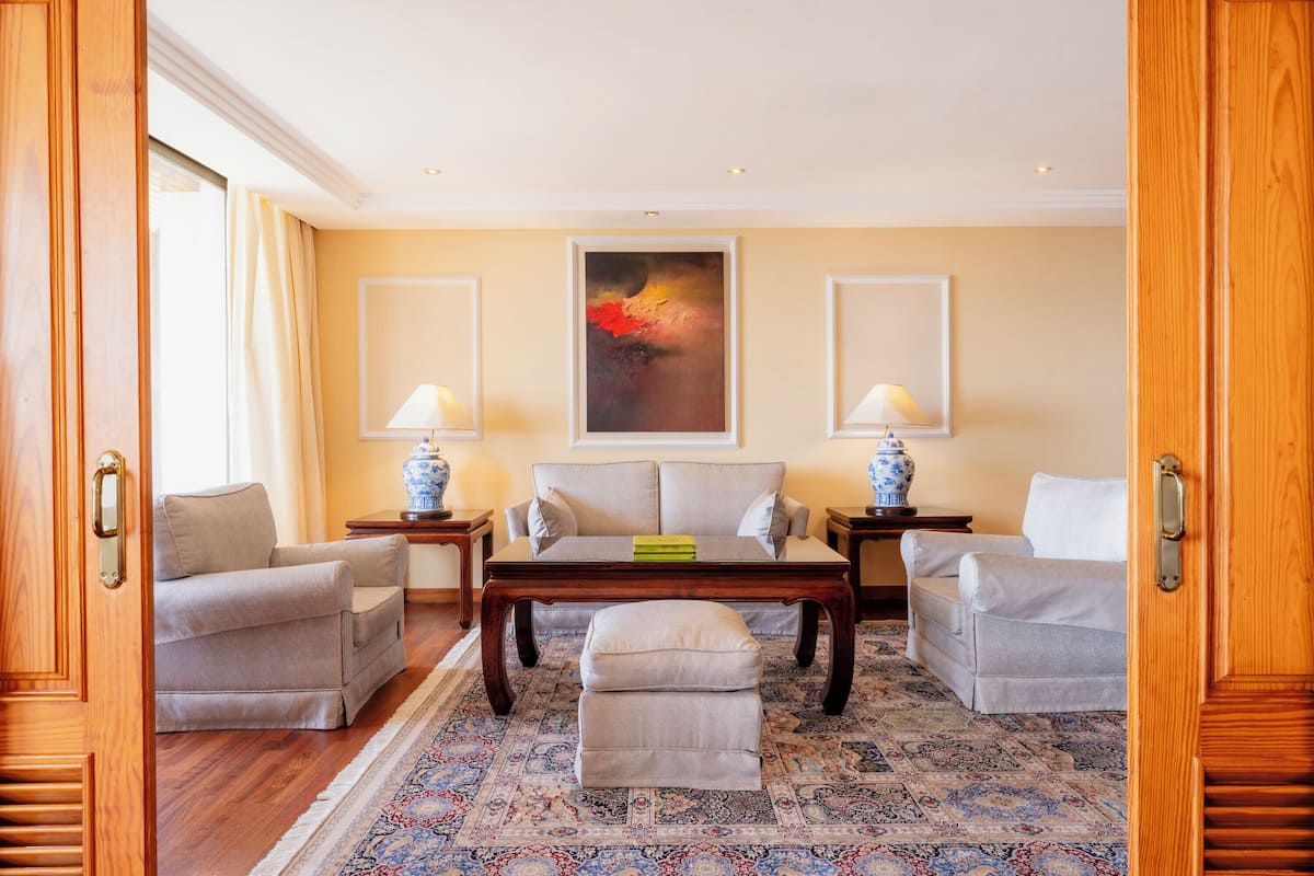 A room with a sofa set, a coffee table, and two lamps. There is a painting on the wall and a patterned rug on the floor.