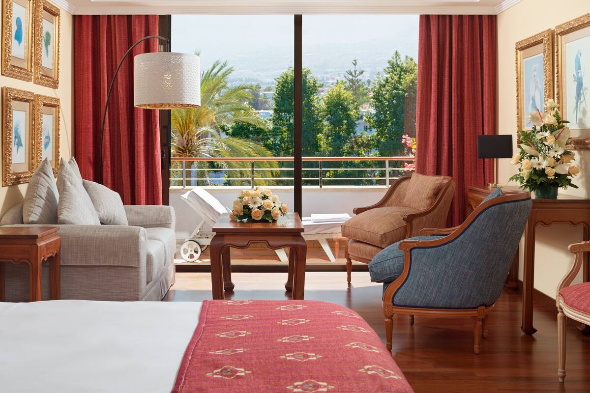 A hotel room with a large bed, two armchairs, a sofa, a small table with a vase of flowers, and a view of the outdoors through large windows.