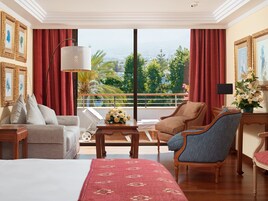 Habitación de hotel con una cama grande, dos sillones, un sofá, una mesita con un jarrón de flores y vistas al exterior a través de amplias ventanas.