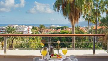 A dining table with a white tablecloth, two wicker chairs, a flower vase, and a fruit plate with drinks on a balcony overlooking a coastal cityscape.