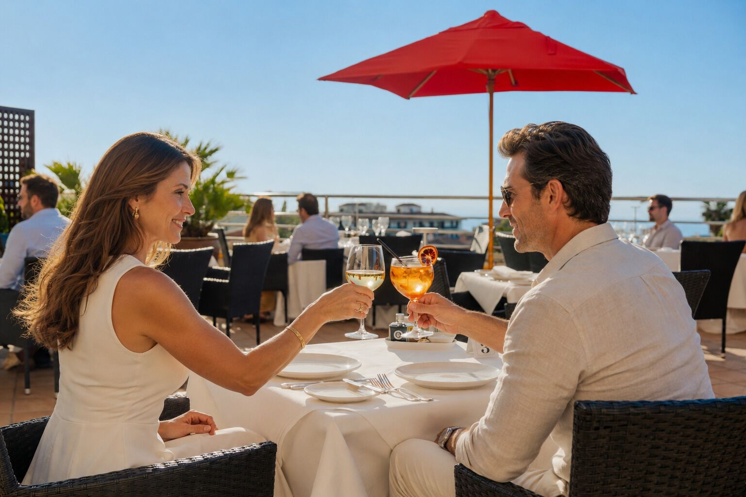 A couple dining outdoors at a restaurant with a red umbrella, white tablecloths, and a view of the sea.