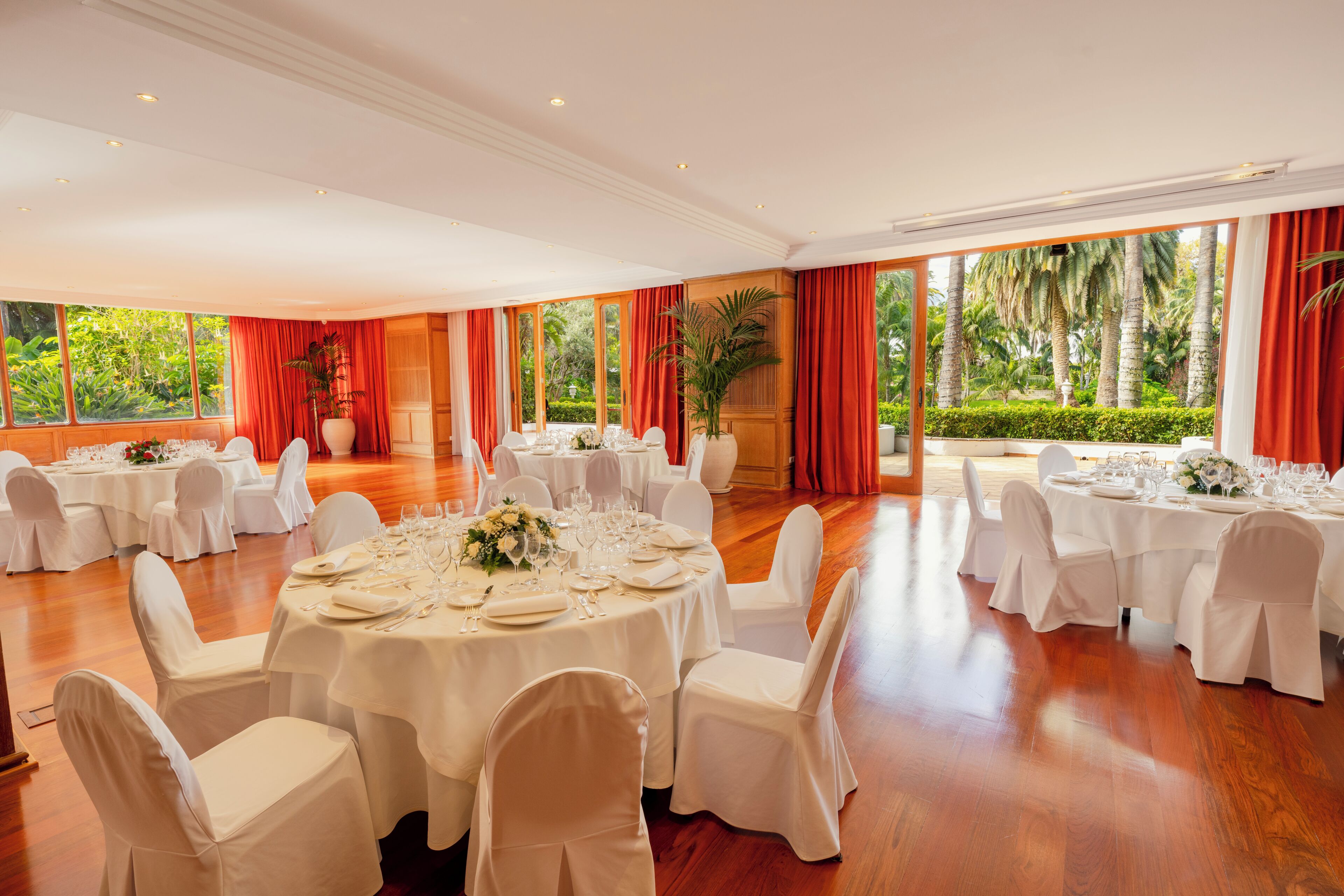 Un salón de banquetes con mesas redondas, sillas blancas y cortinas rojas.