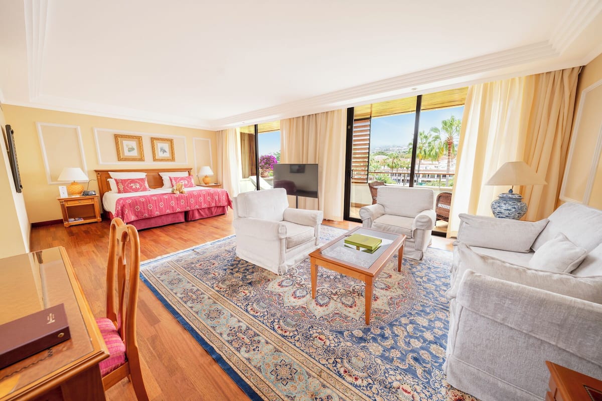 A hotel room with a bed, two armchairs, a sofa, a coffee table, a TV, and a balcony with a view.