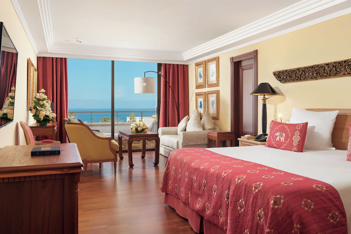 A hotel room with a large bed, a desk, a chair, a small table, and a view of the ocean.