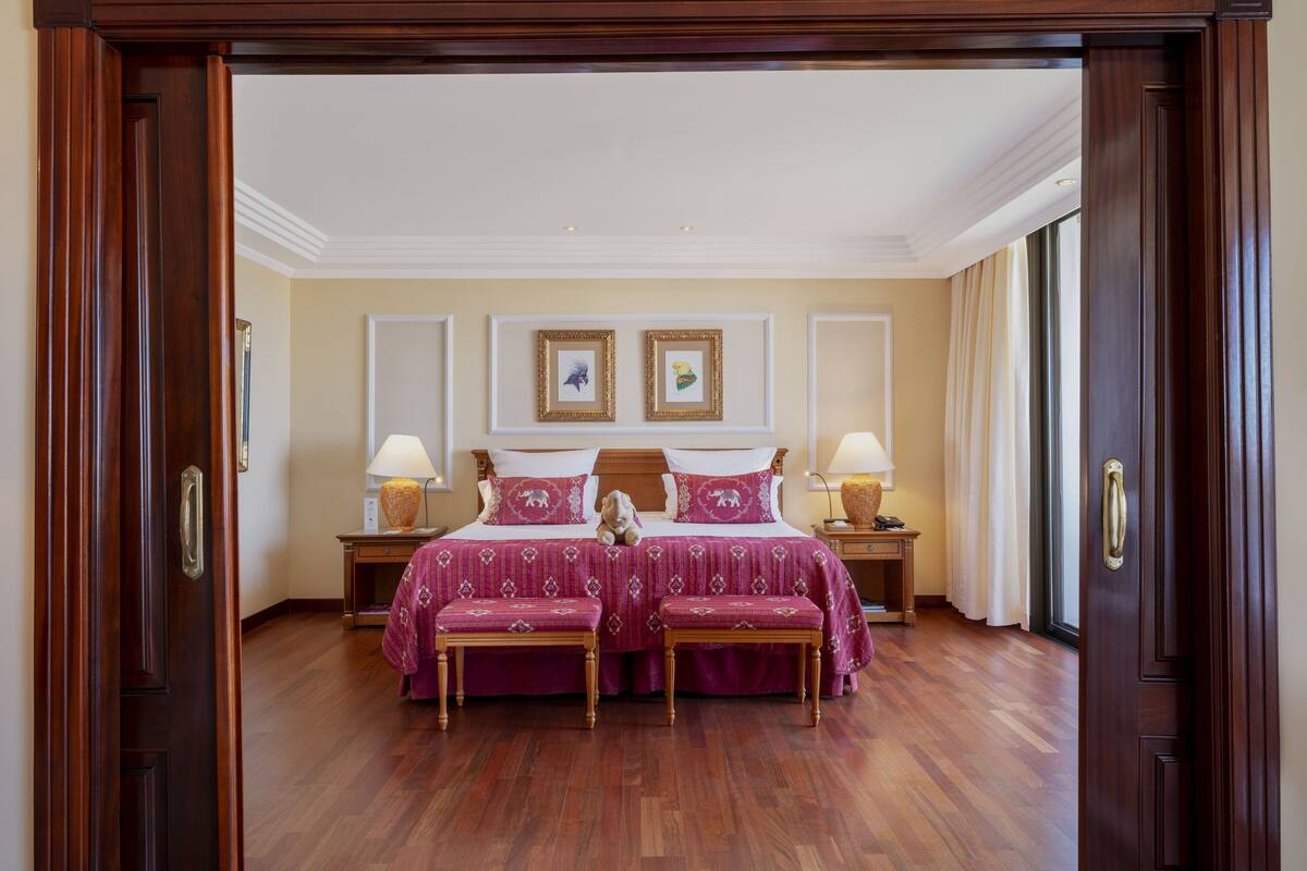 A hotel room with a double bed, two bedside tables with lamps, a wooden bench, and a large window with curtains.