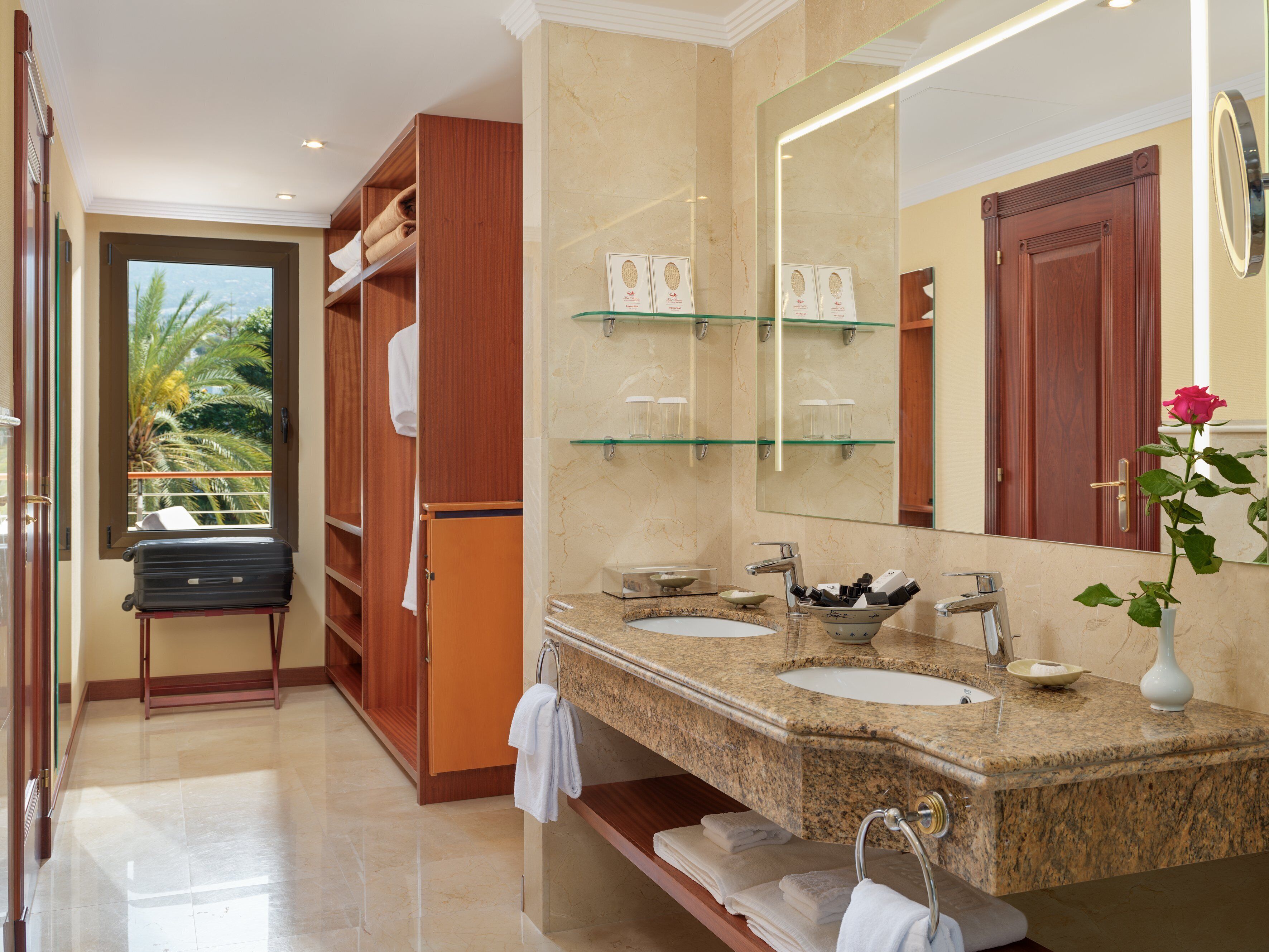 Un baño moderno con lavamanos dobles, un espejo grande y vistas a palmeras a través de una puerta.