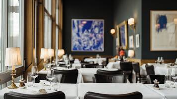 A restaurant with tables set for dining, featuring a modern interior with artwork on the walls and a view of the city through large windows.