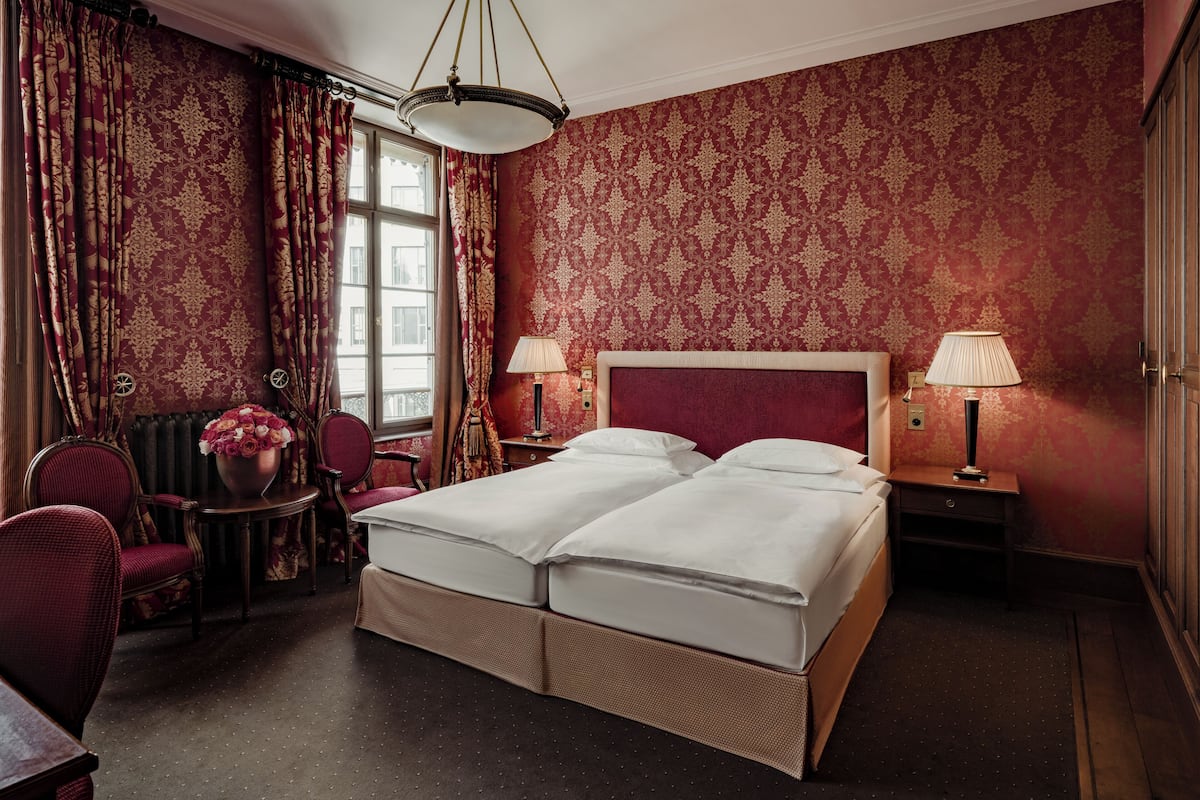 A hotel room with a large bed, two bedside tables, a chair, a small table with a vase of flowers, and a window with curtains.