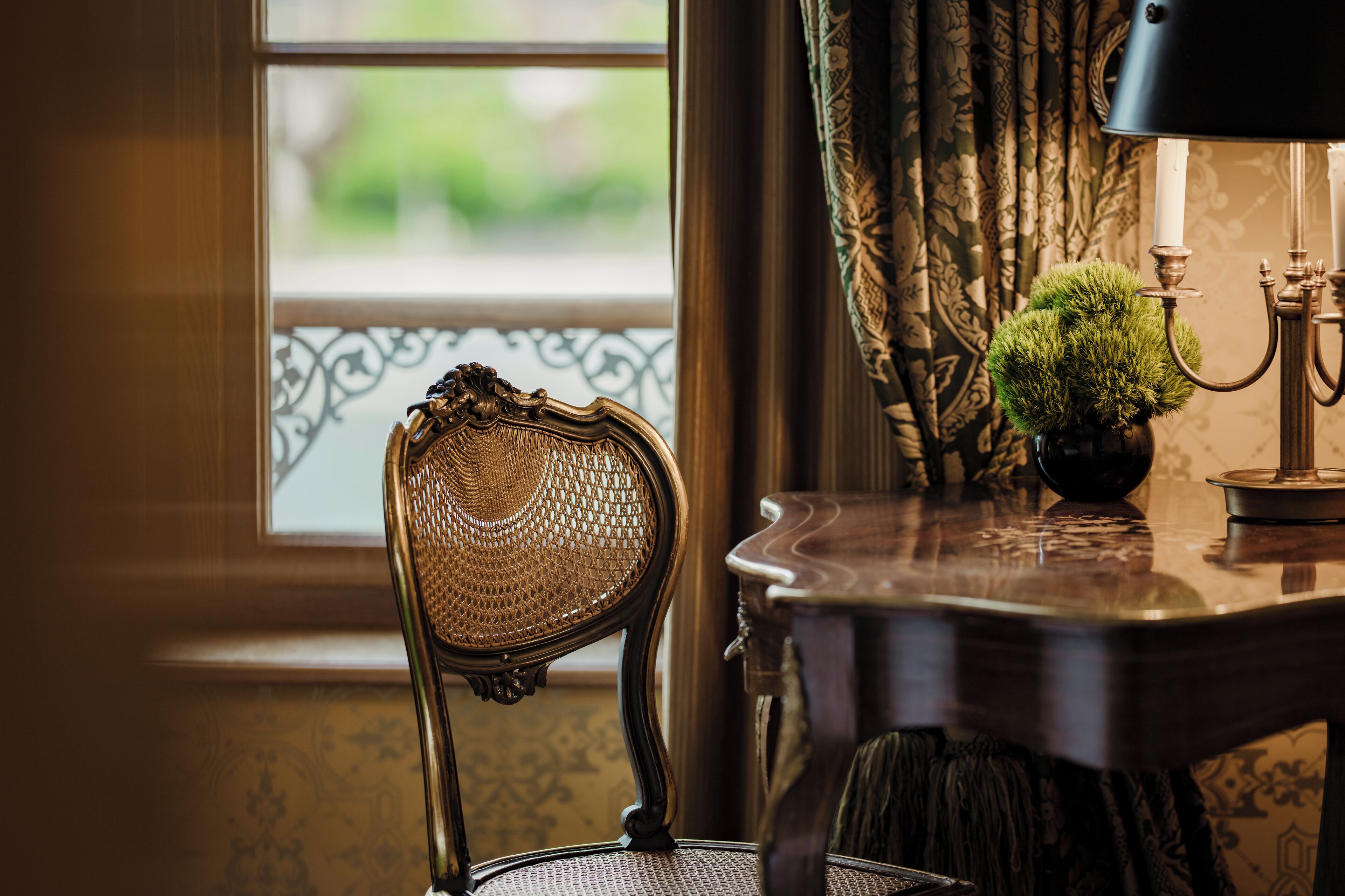 Uma cadeira antiga com encosto trançado, uma mesa com um objeto esférico verde e um candelabro.