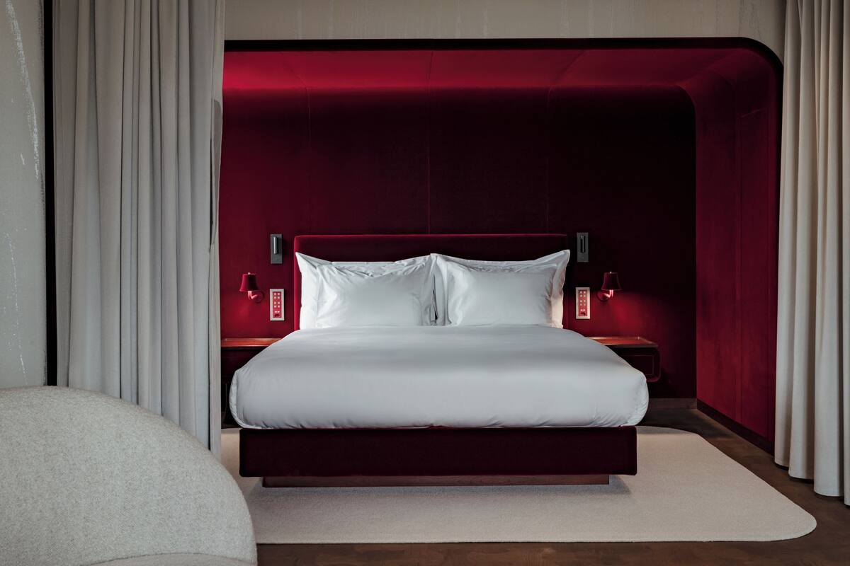 A modern hotel room with a large bed, red accent walls, and a neutral-toned rug.