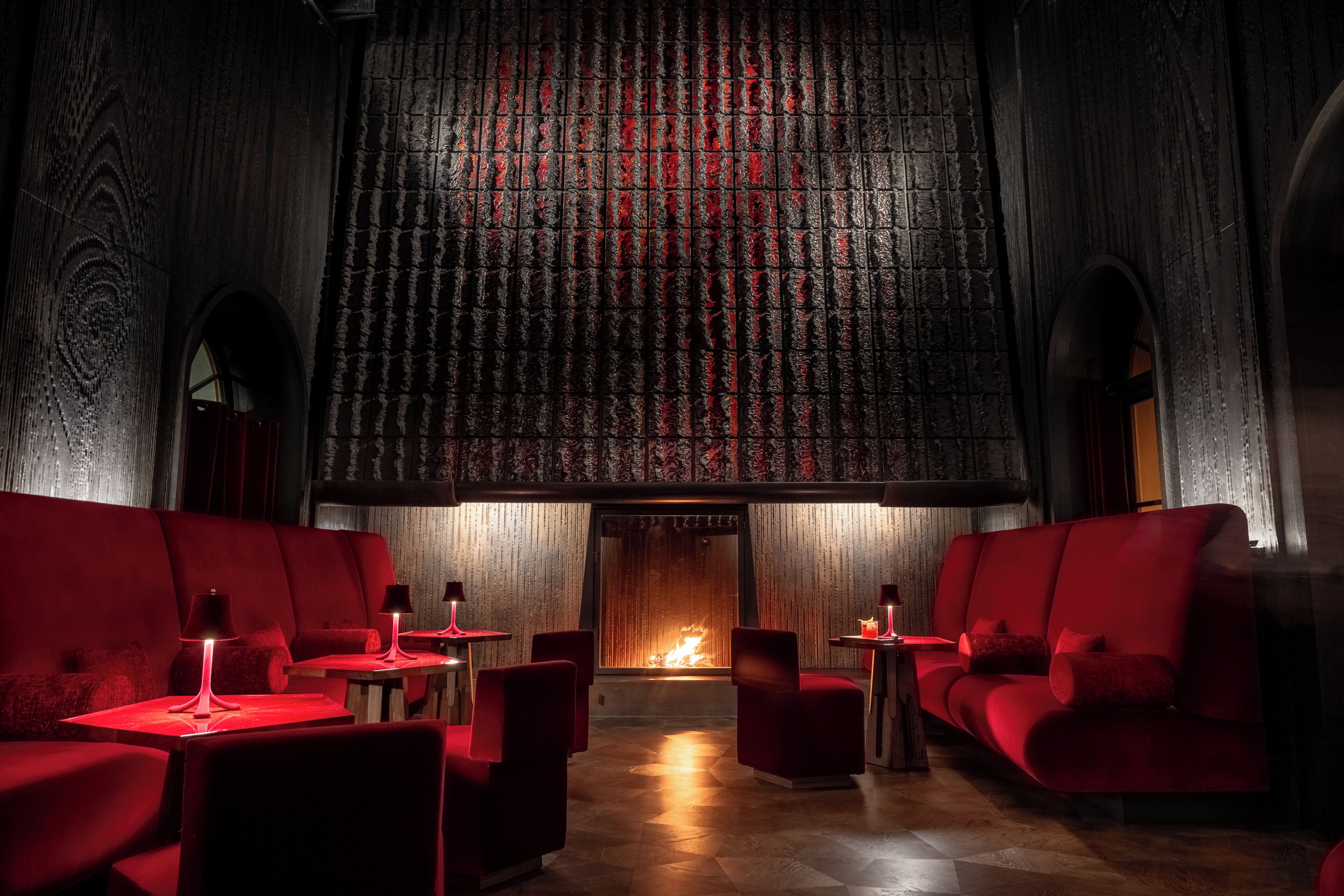 A dimly lit room with red sofas and tables, a patterned wall, and a fireplace.