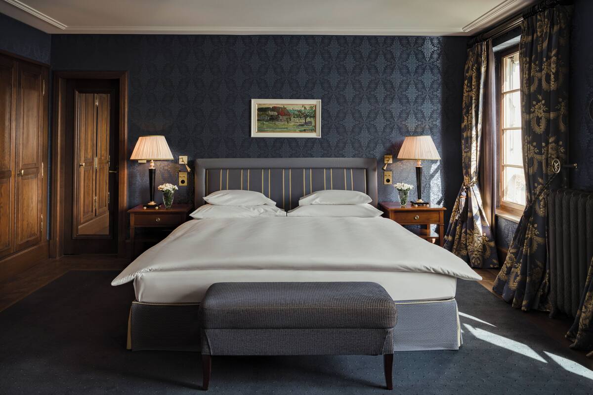 A large bed with a headboard, two bedside tables with lamps, a bench, and a painting on the wall.