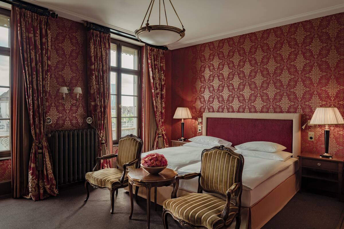 A hotel room with a large bed, two bedside lamps, a small table with a bowl of flowers, two upholstered chairs, and a view of the city through the window.