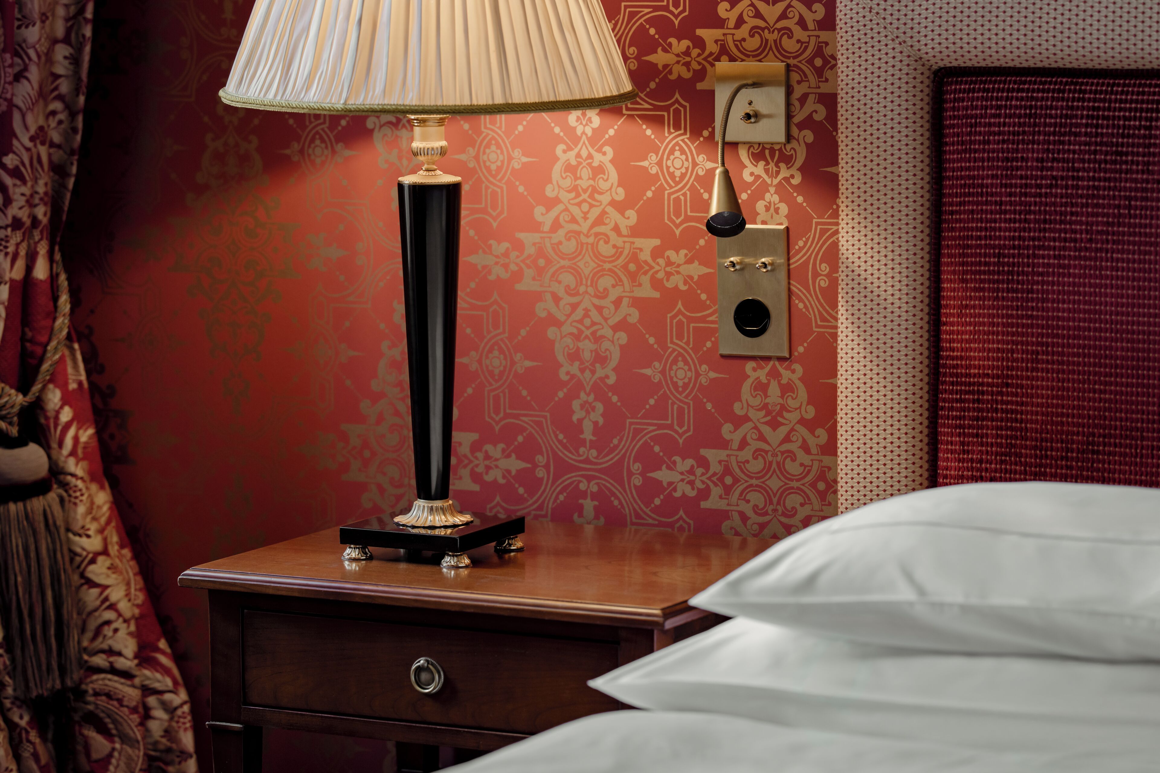 Un lit bien fait, avec du linge de lit blanc, une lampe de chevet, une table de chevet en bois, une horloge murale et un mur décoré d’un motif rouge.