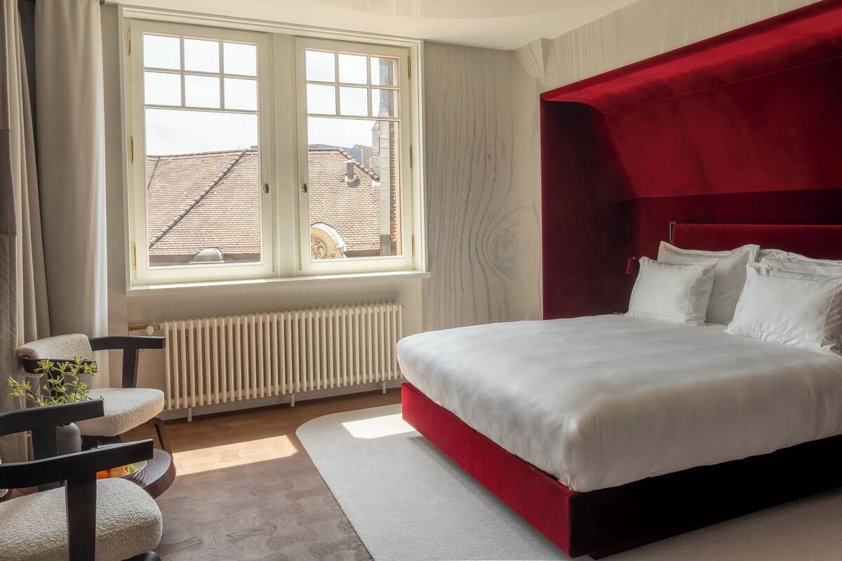 A bedroom with a large bed, a window, a radiator, a chair, and a small table.