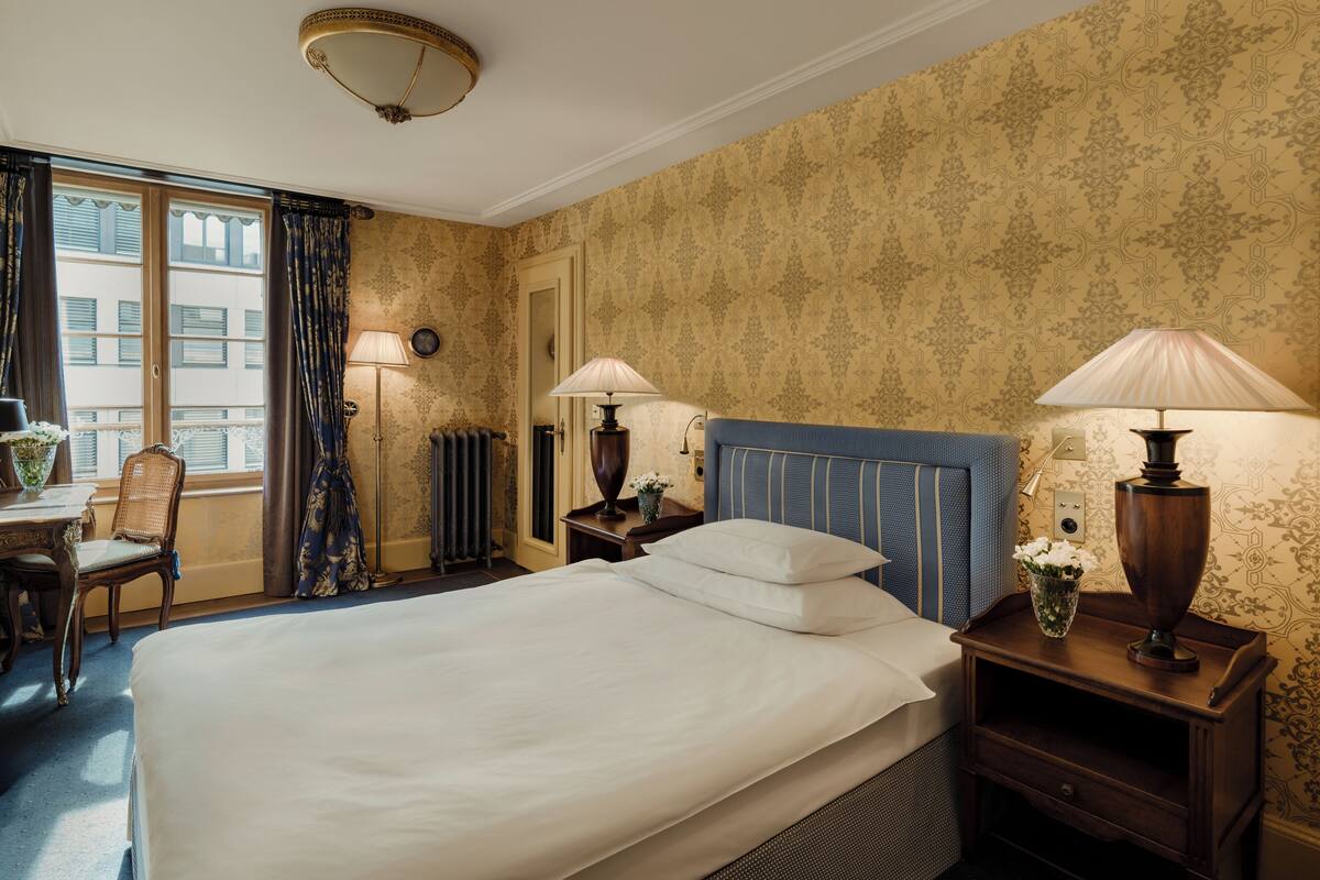 A hotel room with a large bed, two bedside tables with lamps, a chair, a small table with a vase, and a window with curtains.