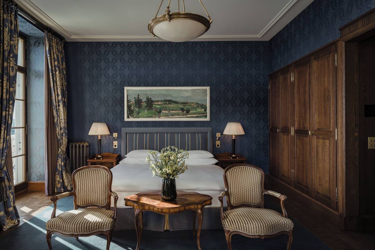 A bedroom with a large bed, two bedside lamps, a painting on the wall, a wooden wardrobe, and two upholstered chairs.
