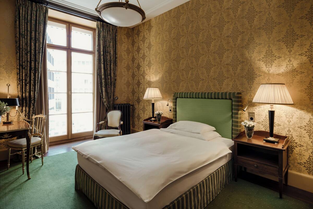 A hotel room with a large bed, two bedside tables with lamps, a chair, a desk, and a large window with curtains.