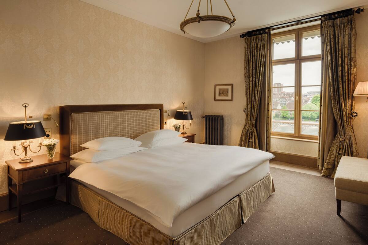 A hotel room with a large bed, two bedside tables with lamps, a chair, a window with curtains, and a radiator.