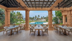 An outdoor dining area with tables and chairs, a pool, and palm trees in the background. - The St. Regis Goa Resort (Cavelossim)