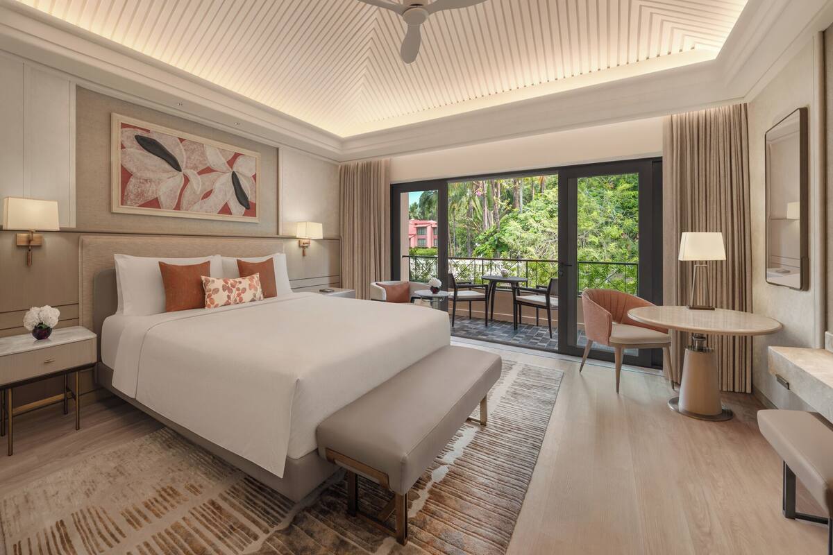 Une chambre d’hôtel spacieuse, dotée d’un grand lit, d’un balcon avec une table et des chaises, et offrant une vue sur la verdure.