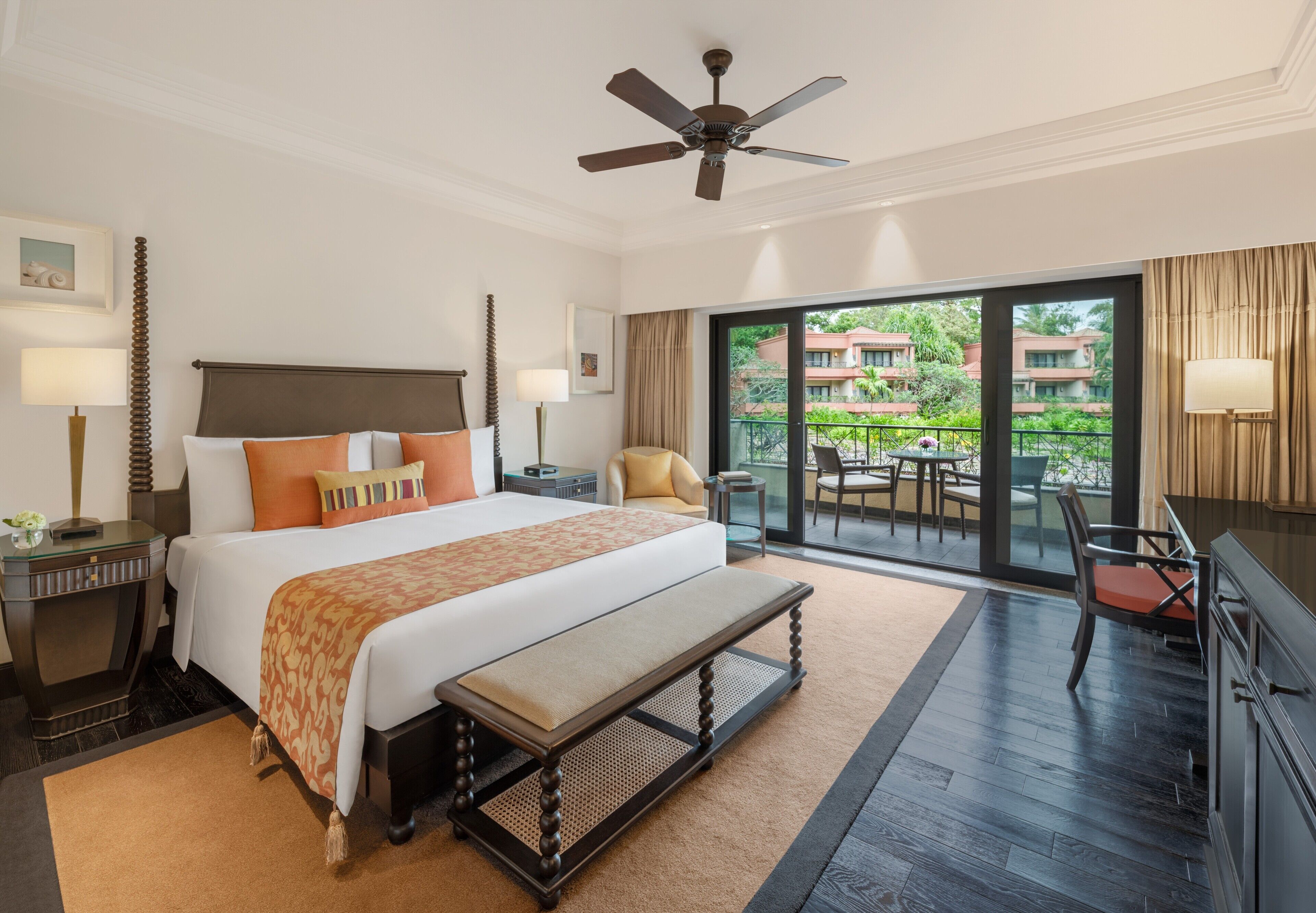 A hotel room with a large bed, a ceiling fan, a balcony with a table and chairs, and a view of greenery outside.