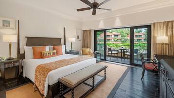 A hotel room with a large bed, a ceiling fan, a balcony with a table and chairs, and a view of greenery outside.