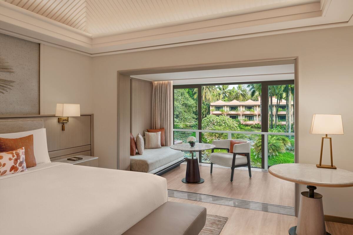 Une chambre d’hôtel avec un lit, un canapé, une petite table et une chaise. Il y a un balcon avec vue sur des palmiers et un immeuble.