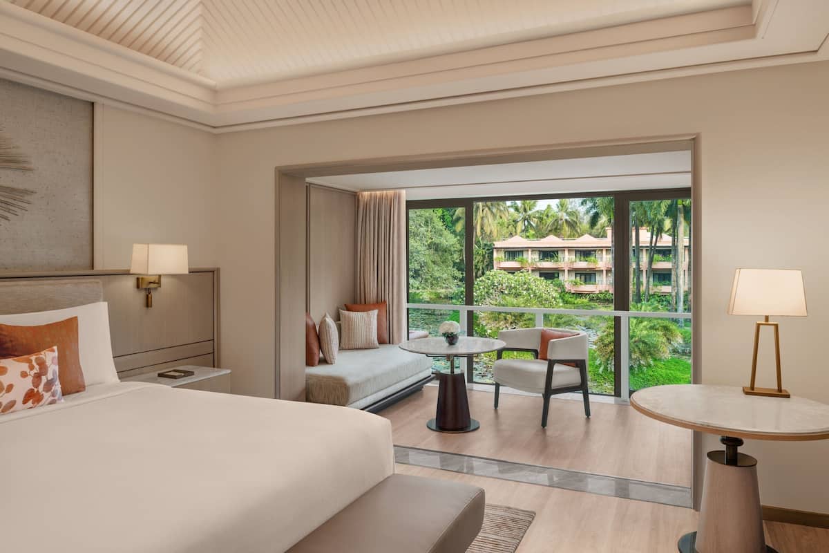 A hotel room with a bed, a sofa, a small table, and a chair. There is a balcony with a view of palm trees and a building.