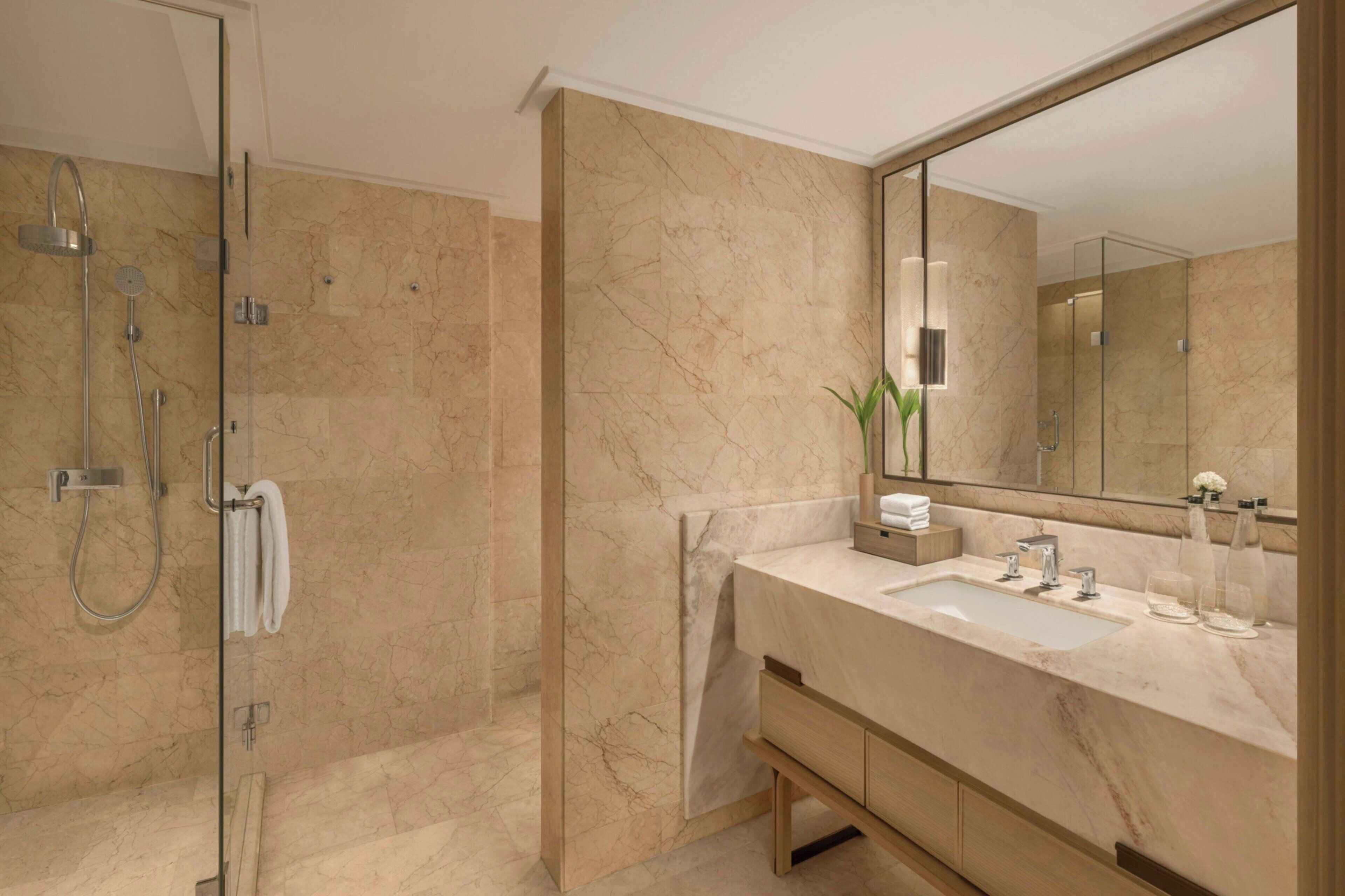 Un baño moderno con ducha envuelta en vidrio, pared de azulejos de mármol, un espejo grande y un mueble de tocador de madera con lavamanos.