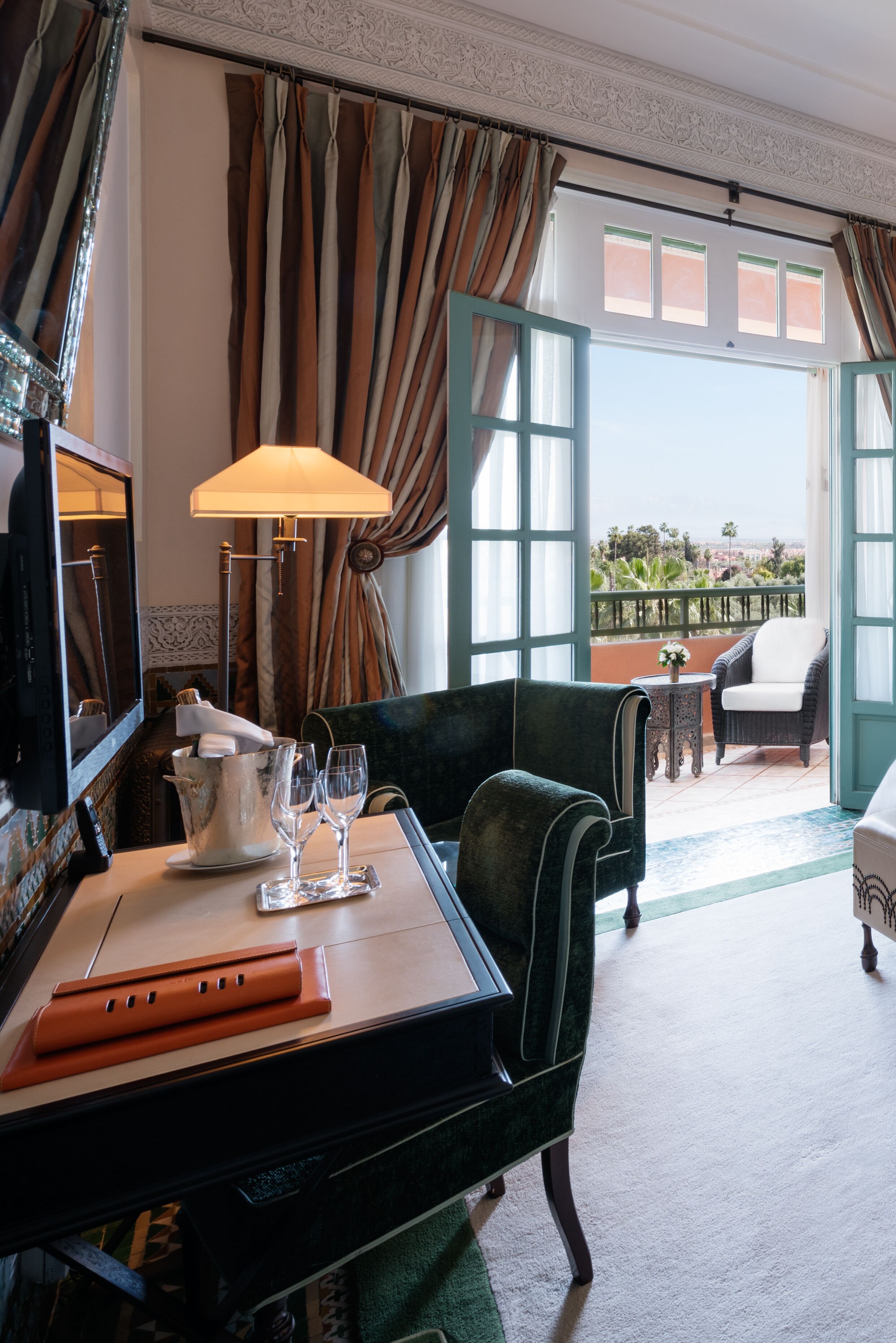 Quarto de hotel com varanda, uma mesa com abajur, uma cadeira e vista para o exterior.