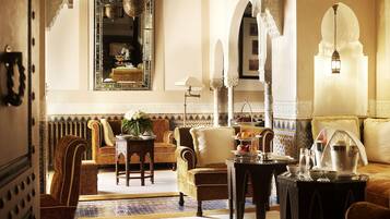 A traditional interior with ornate decor, a seating area with sofas and armchairs, a dining table with a pitcher and glasses, and a large mirror.