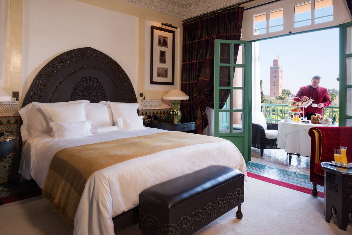 Camera d'hotel con un letto grande, una panca, un'area pranzo con tavolo e sedie, e vista su una torre attraverso una porta aperta.