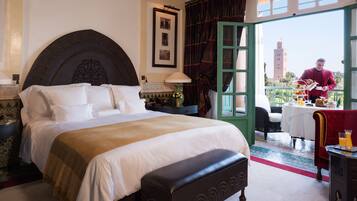 A hotel room with a large bed, a bench, a dining area with a table and chairs, and a view of a tower through an open door.