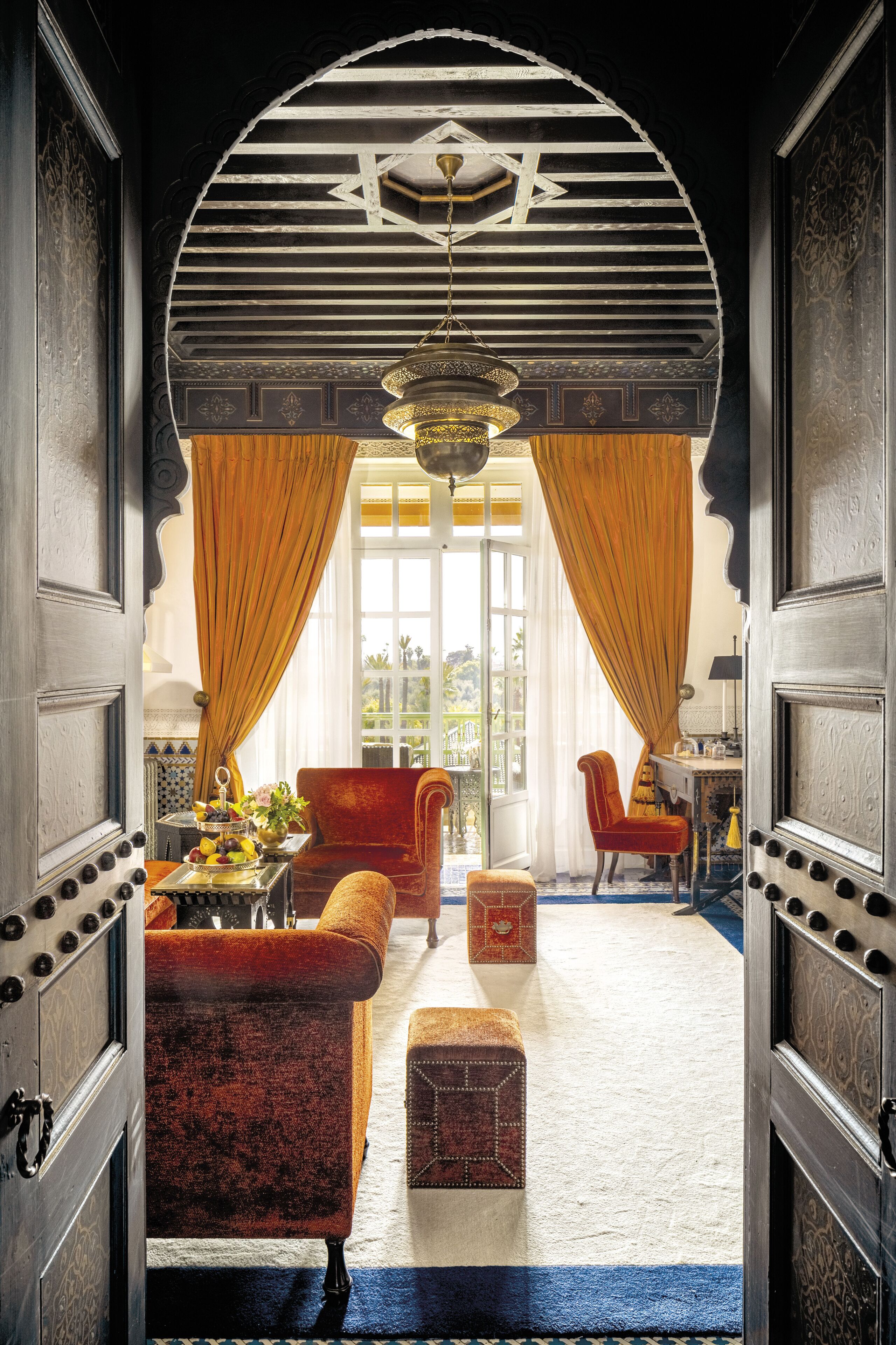Une porte ouverte donne sur une pièce où se trouve un canapé en velours rouge, une petite table avec un vase, et d’où l’on aperçoit un balcon.