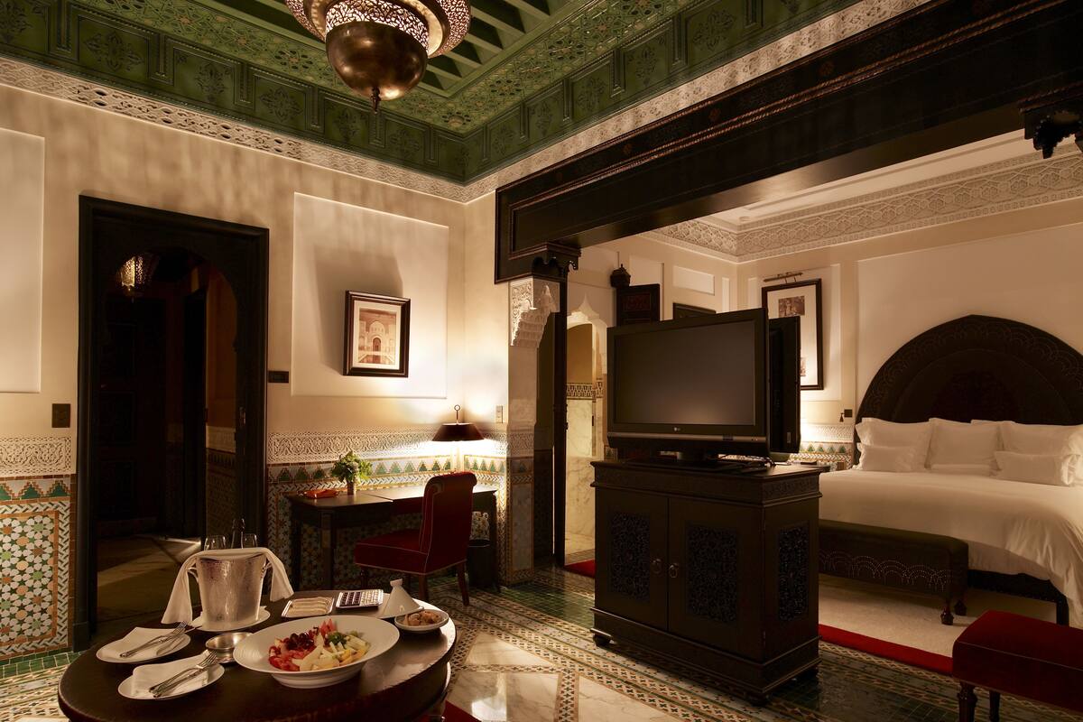 Camera d'albergo con un letto, televisione, zona pranzo con tavolo e sedie, e un soffitto decorativo.