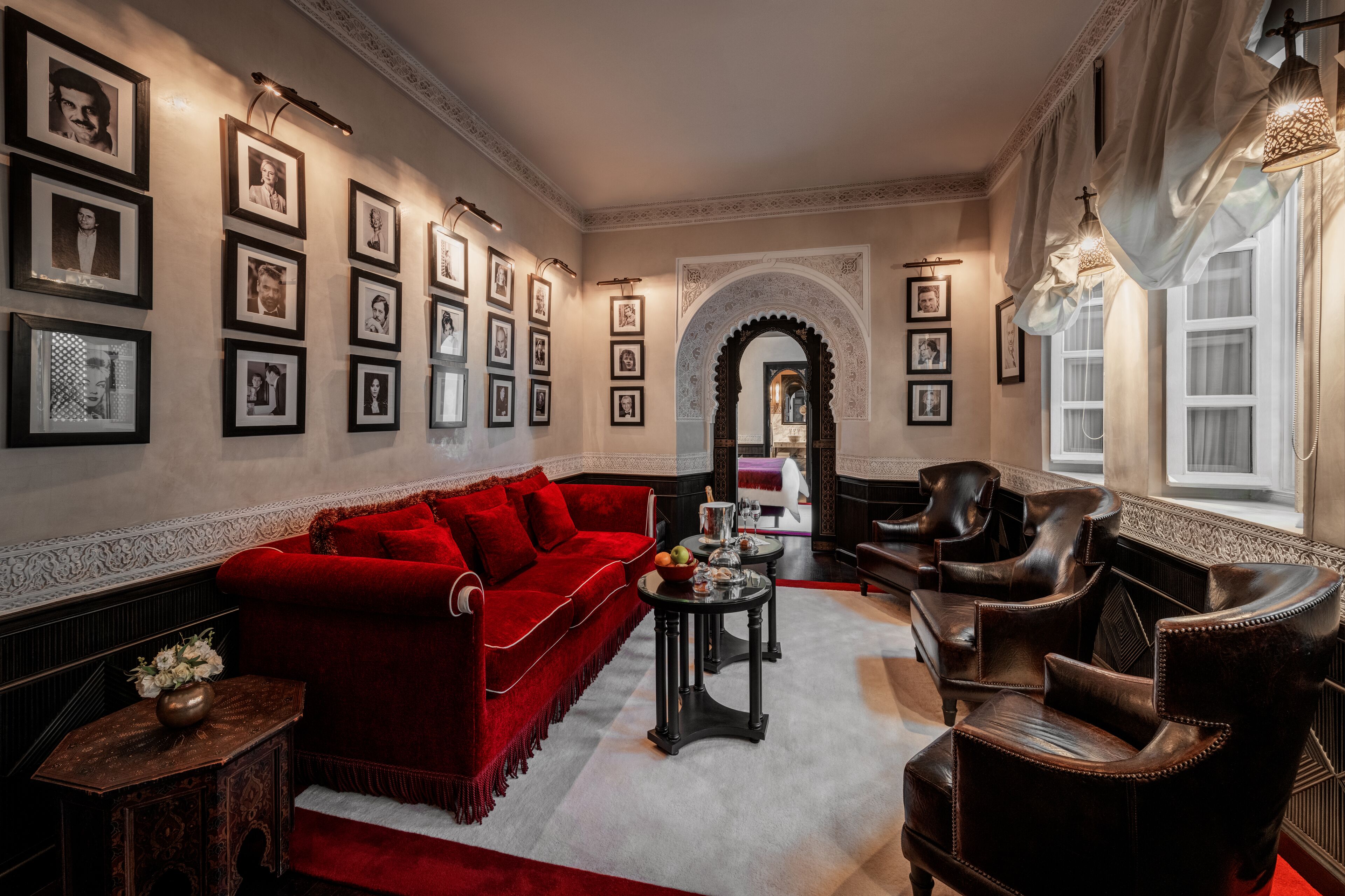 A spacious living room with a red sofa, leather armchairs, and a coffee table. There are framed pictures on the walls and a decorative archway.