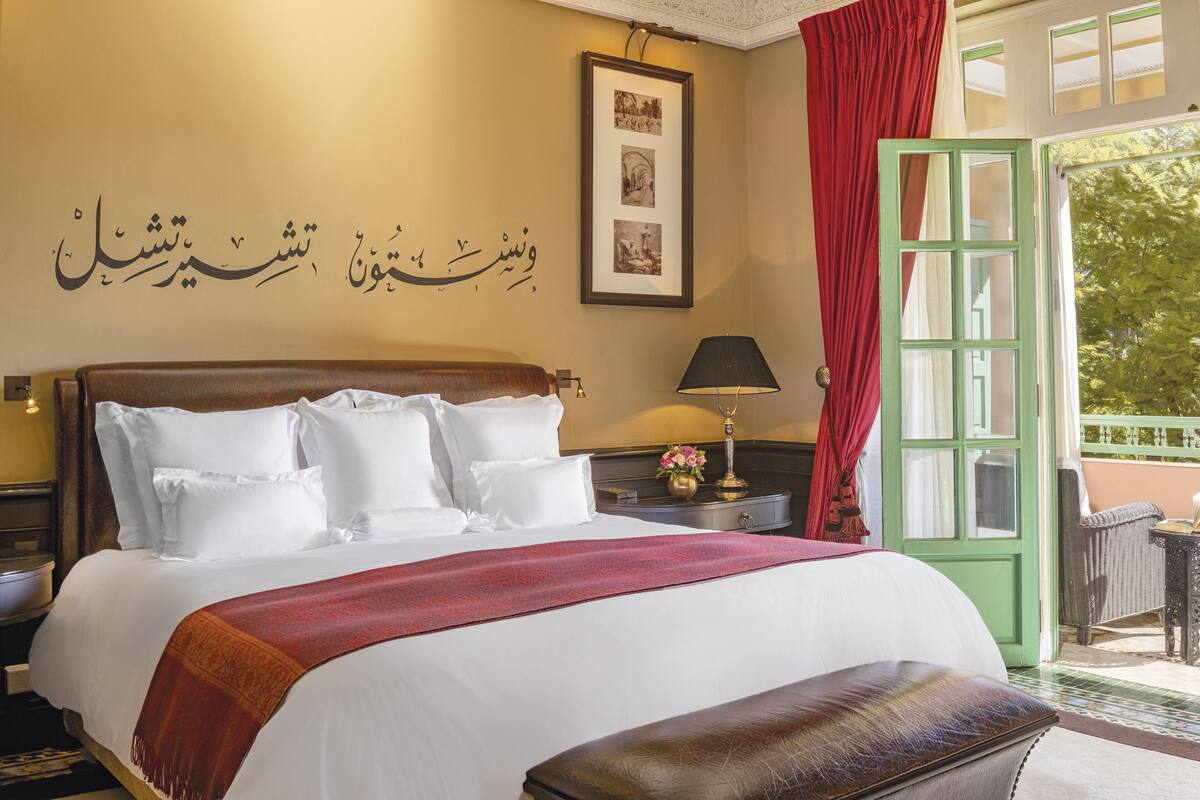 Una camera d'hotel con un letto grande, un comodino, una lampada, una finestra con tende rosse e un muro con calligrafia araba.