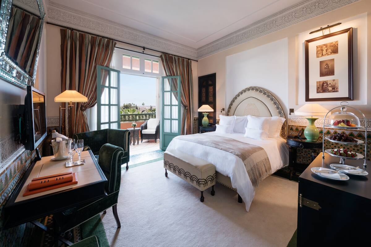 Una camera d'albergo con un letto grande, una scrivania con una lampada, una sedia, un tavolo da pranzo con un cesto di frutta e vista sull'esterno.