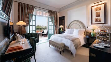 Une chambre d’hôtel avec un grand lit, un bureau avec une lampe, une chaise, une table à manger agrémentée d’un présentoir de fruits et une vue sur l’extérieur.