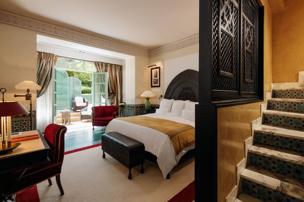 Una camera d'albergo con un letto grande, una scrivania con una lampada, una sedia rossa e una panca. Si vede una scala con ringhiera decorata e uno scorcio di un giardino attraverso una porta aperta.