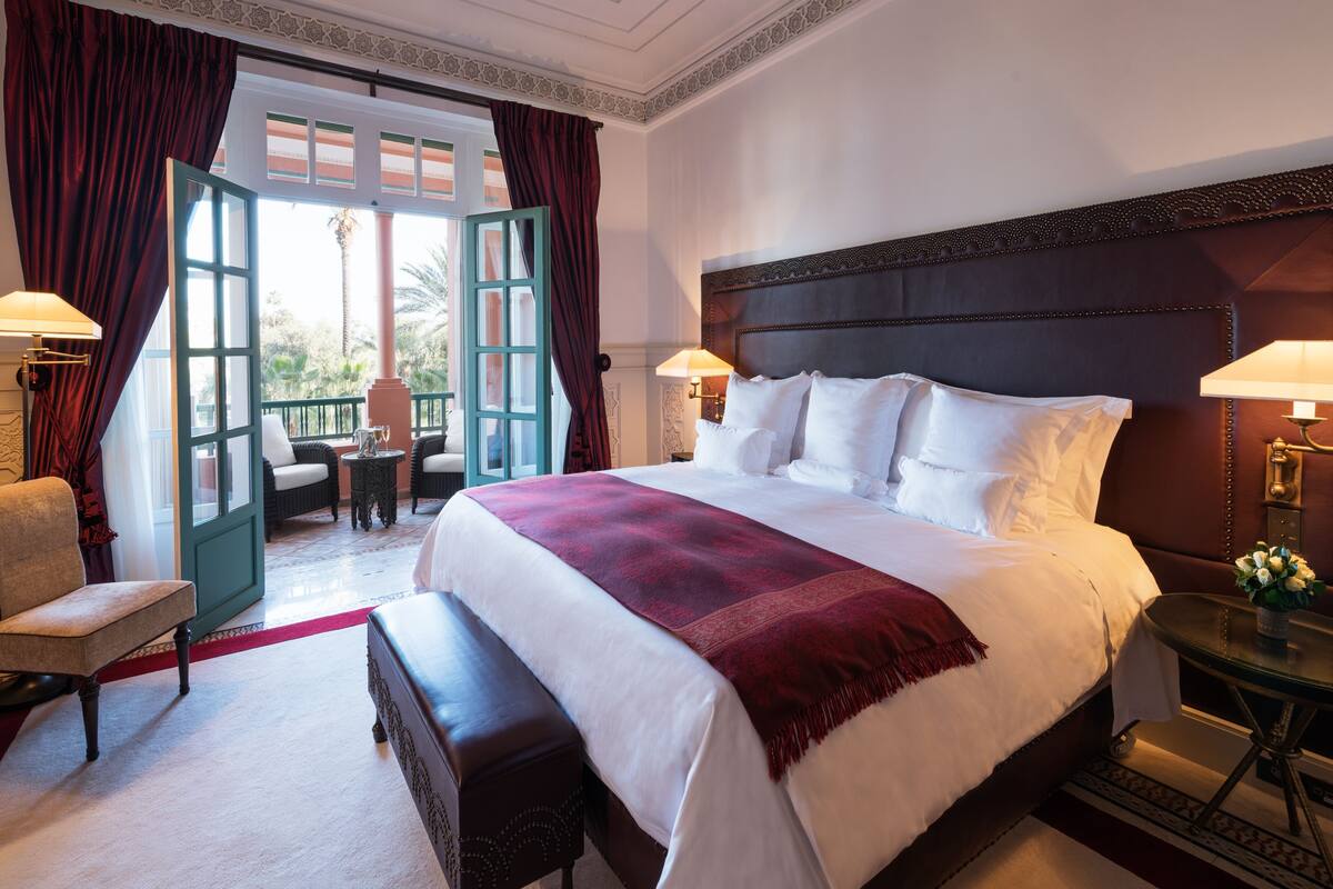 Un letto grande con lenzuola bianche e una coperta rossa, una testiera in legno, due lampade sui comodini, una panca e vista all'esterno attraverso una porta aperta.