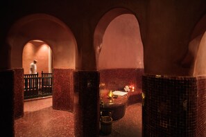 A dimly lit room with arched ceilings and tiled walls, featuring a bench with a candle and a small table with a book and a cup.