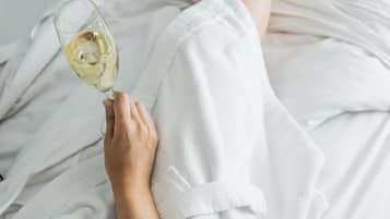 A person lying in bed with a glass of white wine, wearing white bathrobe.
